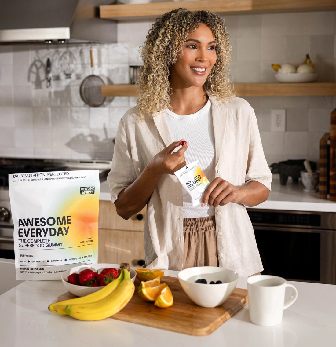 Woman holding Awesome Everyday gummies in a kitchen with fruit and a mug on the counter.