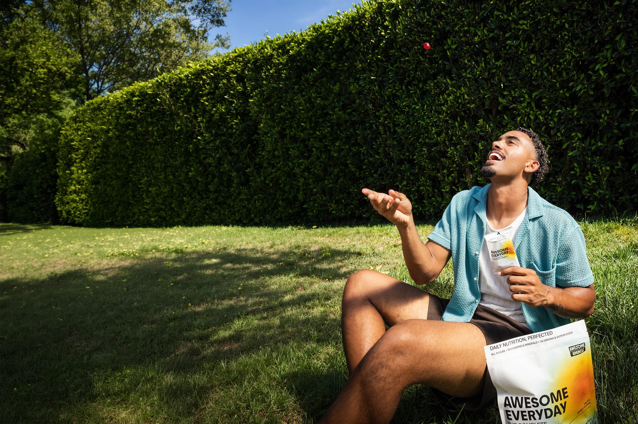 A man sitting on the grass tossing a gummy into his mouth, a pouch of Awesome Everyday beside him
